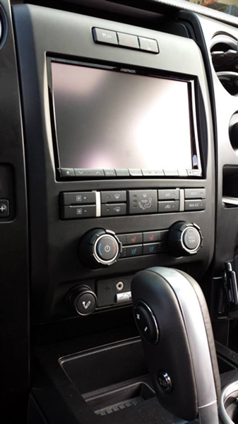 Center console of a vehicle featuring a large touchscreen, control buttons, and knobs, with a gear shift lever visible at the bottom.