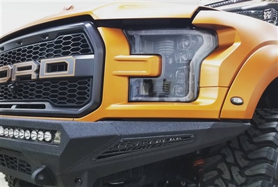 Close-up of the front of an orange Ford truck, highlighting the grille with the "FORD" emblem, headlight, and a rugged front bumper with additional lights.