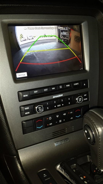 Vehicle dashboard showing a rearview camera display with parking guidelines, control buttons and knobs below, and part of the gear shift lever visible.