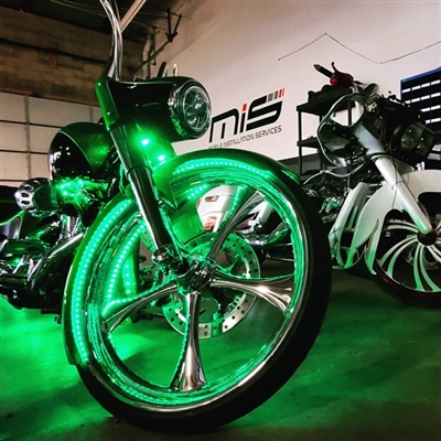A close-up view of a motorcycle with large, chrome wheels illuminated by green lights, parked indoors next to another motorcycle.