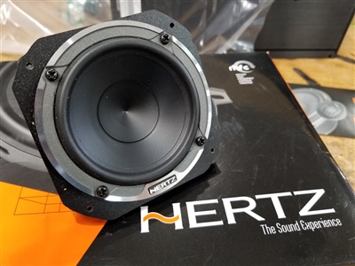 A close-up view of a Hertz car speaker placed on top of its packaging box, labeled "Hertz The Sound Experience."