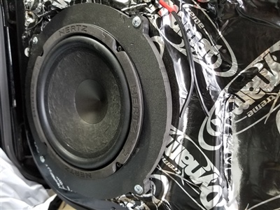 A close-up view of a car speaker installed on a door panel, with soundproofing material labeled "Dynamat" surrounding it.