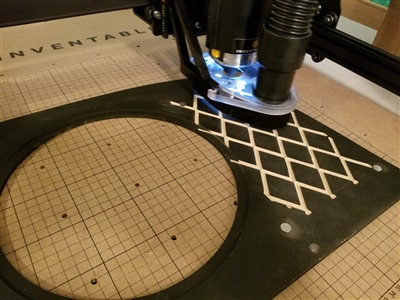 A close-up view of a CNC machine cutting a geometric pattern into a material, with a large circular cutout already made, on a worktable marked with a grid.