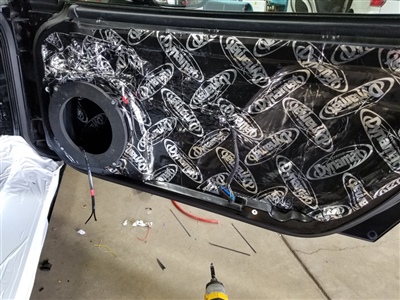 The interior of a car door panel with a black speaker installed and soundproofing material labeled "Dynamat" covering the entire surface.