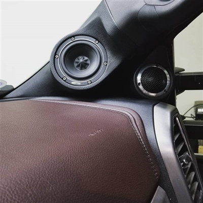 A close-up view of a car's interior showing a round speaker installed on the A-pillar and a portion of the dashboard with brown leather trim.