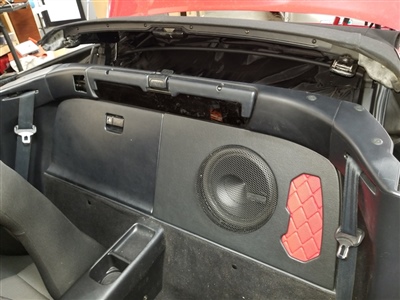 The interior of a car with the rear seat removed, revealing a custom audio setup with a subwoofer installed in the back panel and a red mesh accent on the side.