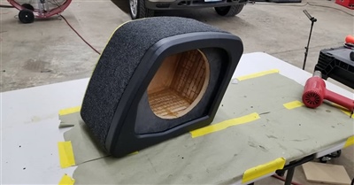 A custom-made, carpeted subwoofer enclosure with an unfinished circular cutout, displayed on a workbench in a garage or workshop.