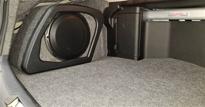  The interior of a car trunk with a custom subwoofer enclosure installed in the corner, surrounded by carpeted lining and adjacent to other equipment.