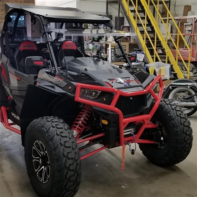 A black and red off-road vehicle with large tires and rugged suspension parked inside a workshop, with yellow stairs and various equipment in the background.