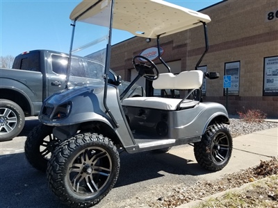 A customized golf cart with large off-road tires and a lifted suspension, parked outside a brick building.