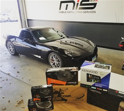 Black sports car parked inside a garage with "Mobile Installation Services" signage on the wall in the background, surrounded by various car audio and electronics boxes.