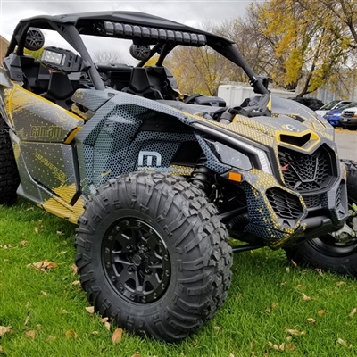 A close-up view of a black and yellow off-road vehicle with large rugged tires, parked on a grassy area with trees and other vehicles in the background.