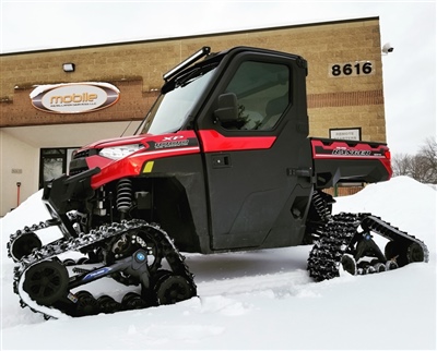 A red and black off-road vehicle equipped with snow tracks instead of wheels, parked outside a building on a snowy day.