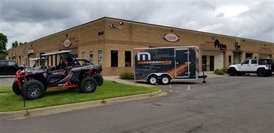 An exterior view of a brick building with an off-road vehicle, a trailer with "MS" branding, and a white Jeep parked outside.