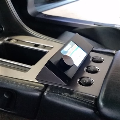 Close-up of a vehicle's center console featuring a small display screen with a control knob and three buttons, surrounded by metallic and black leather trim.