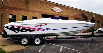 A sleek white speedboat with purple and pink accents on a trailer, parked in front of a brick building.