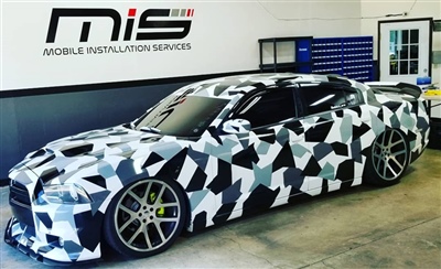 Modern car with a black, white, and grey camouflage wrap, parked inside a garage with "Mobile Installation Services" signage on the wall in the background.