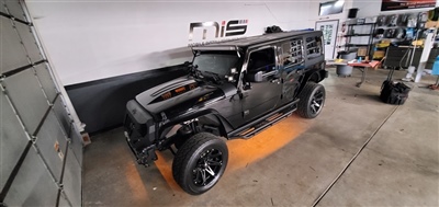 A black customized Jeep with large wheels and an open hood, parked inside a garage with "Mobile Installation Services" sign on the wall.