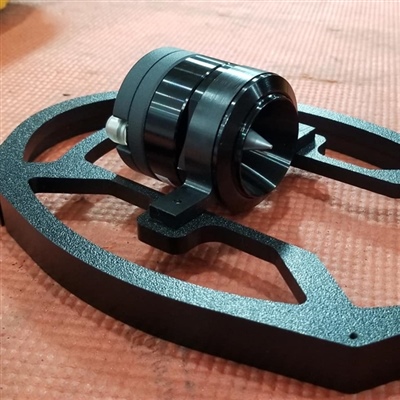 Black mechanical component with a cylindrical central part and a flat, circular base, placed on a textured red surface.