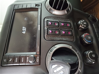 Close-up of a car dashboard featuring a large touchscreen display, control buttons with pink backlighting, air vents, and climate control knobs.