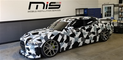 A sports car with a black and white geometric camouflage wrap, parked inside a garage with "Mobile Installation Services" sign on the wall.