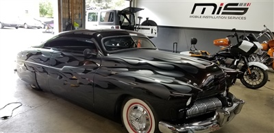 A shiny black vintage car with whitewall tires parked inside a garage, with motorcycles and a "Mobile Installation Services" sign in the background.