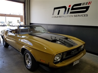 A yellow vintage convertible parked inside a garage with "Mobile Installation Services" sign on the wall in the background.
