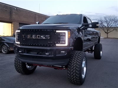 A lifted black truck with large off-road tires and LED lights, parked in front of a brick building at dusk.