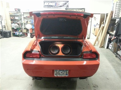 The rear view of an orange car with the trunk open, revealing two large subwoofers installed inside.