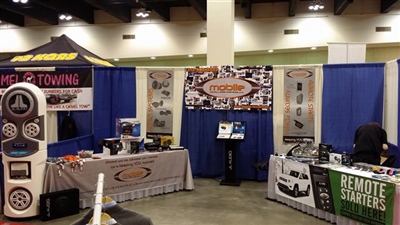 A trade show booth featuring displays of automotive audio equipment, banners, and promotional materials, with blue curtains in the background and tables showcasing products.