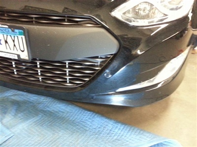 A close-up view of the front bumper and grille of a black car, with part of a blue cloth visible on the ground in front of it.